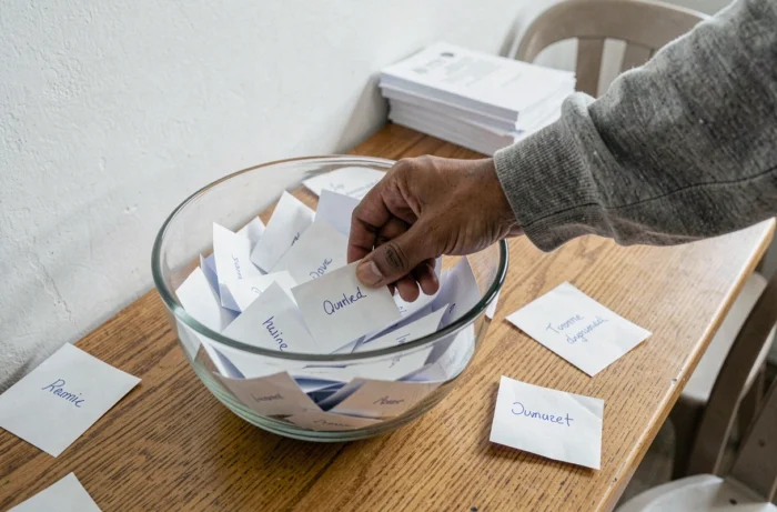 Main piochant un billet dans un tirage au sort citoyen, avec des noms manuscrits visibles sur des papiers dépliés sur une table en bois